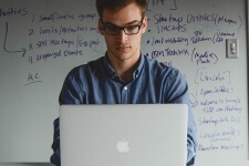 Ein Projektmanager tippt an einem Laptop, im Hintergrund ist ein beschriebenes Whiteboard zu erkennen