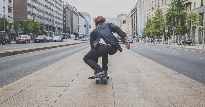 Ein Mann im Anzug skatet einen Buergersteig einer stark befahrenen Strasse entlang