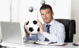 Sportmanager sitzt an Schreibtisch und haelt einen Fussball zwischen den Haenden