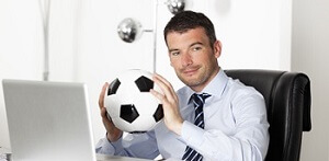 Sportmanager sitzt an Schreibtisch und haelt einen Fussball zwischen den Haenden