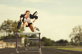 Eine Sportökonomin springt im Businesslook über eine Hürde.